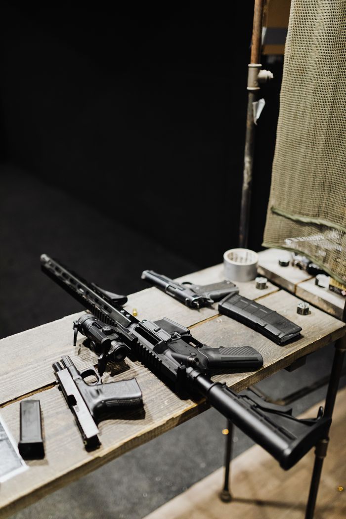 Services Assorted firearms arranged on a wooden table in an indoor shooting range setting.