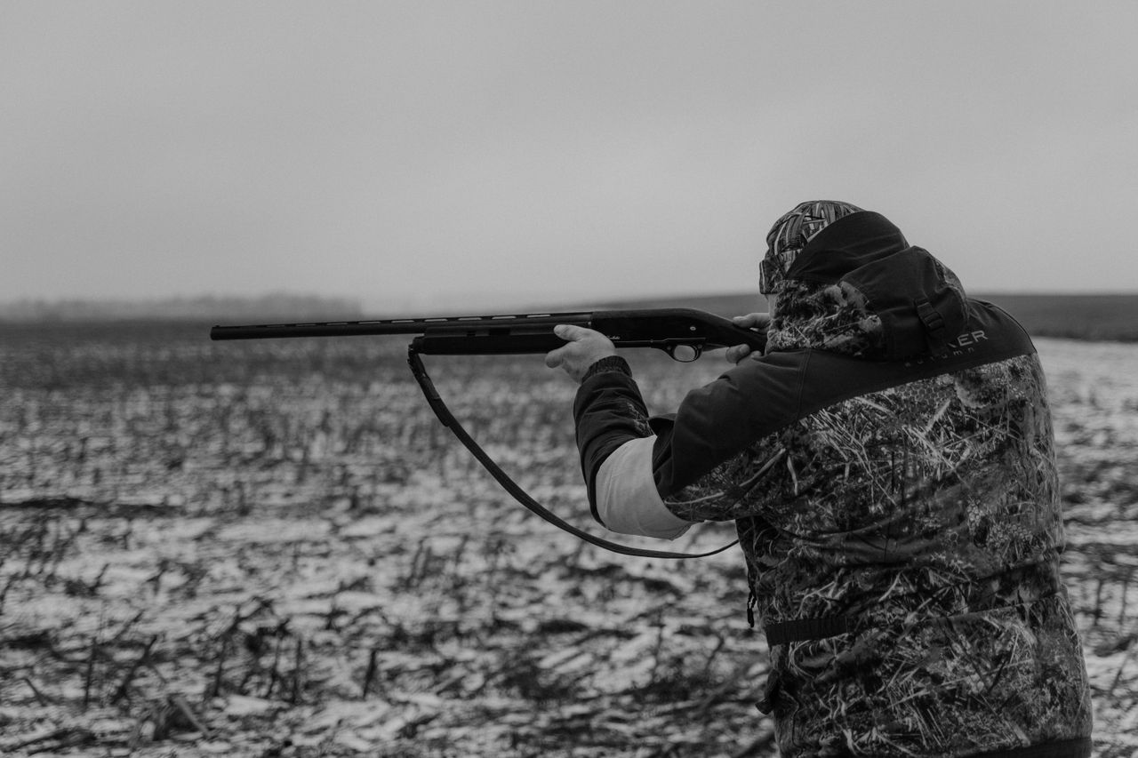 About A hunter in winter attire aims a rifle in a snowy countryside setting.